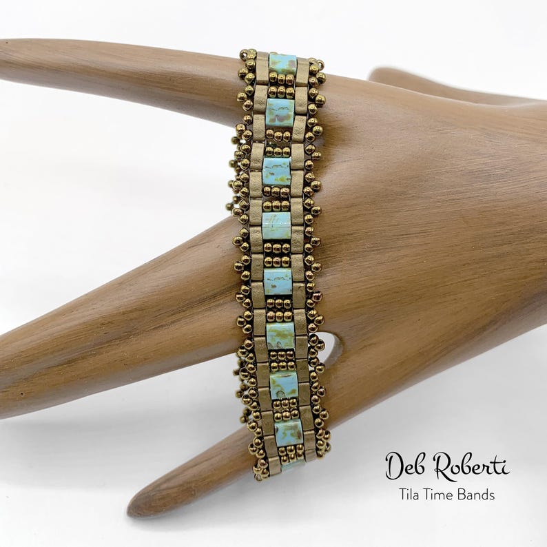 May include: A beaded bracelet with a gold and turquoise color scheme. The bracelet is made with small, square beads and has a delicate, intricate design. The bracelet is on a wooden hand model. The text "Deb Roberti Tila Time Bands" is visible below the bracelet.