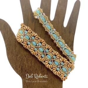 Könnte beinhalten: Zwei Perlenarmbänder, eines mit einem hellblauen und goldenen Muster und das andere mit einem hellgrünen und goldenen Muster, auf einer Holzhand präsentiert. Die Armbänder sind mit "Duo Lace Bracelets" beschriftet.