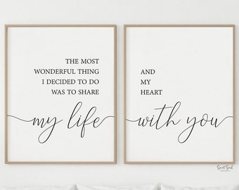 May include: Two framed prints with black calligraphy text on a white background. The text reads "The most wonderful thing I decided to do was to share my life with you". The prints are hanging on a white wall above a white bed with white bedding.