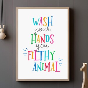 May include: A rectangular wall art print in a light wood frame featuring the colorful text WASH your HANDS you FILTHY ANIMAL. The words are written in a mix of blue, orange, green, pink, and yellow fonts against a plain white background.