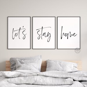 May include: Three black framed prints with white backgrounds and black calligraphy. The prints read "let's", "stay", and "home". The prints are hanging on a white wall above a bed with white bedding.