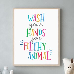 May include: A rectangular wall art print in a light wood frame featuring the text "WASH your HANDS you FILTHY ANIMAL" in a variety of bright, multi-colored letters. The words are stacked vertically against a plain white background.