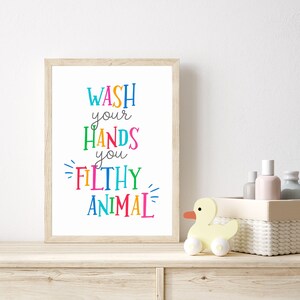 May include: A rectangular framed wall art print featuring the text WASH your HANDS you FILTHY ANIMAL in a variety of bright, multi-colored letters. The frame is made of light-colored wood and the background of the print is solid white.