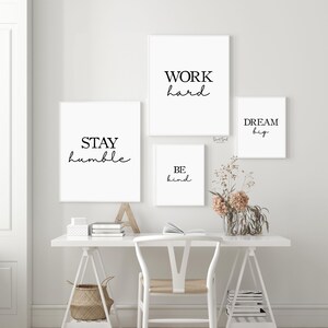 May include: Four framed wall art prints with motivational quotes in black script on white backgrounds. The quotes read "WORK hard", "STAY humble", "BE kind", and "DREAM big". The prints are displayed above a white desk with a chair and decorative items.