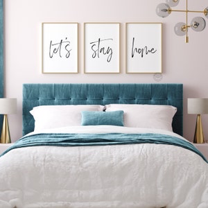 Pu&ograve; includere: Tre stampe incorniciate con le parole "let's", "stay" e "home" in scrittura nera su sfondo bianco. Le stampe sono appese sopra una testata imbottita blu turchese su un letto con lenzuola bianche e una coperta blu turchese.