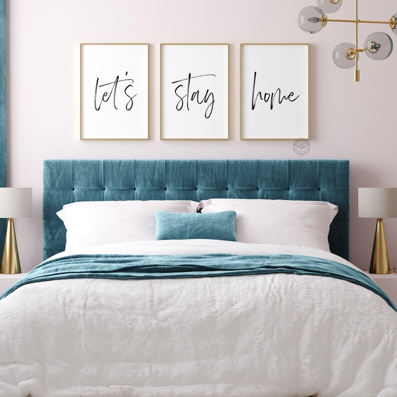 Pu&ograve; includere: Tre stampe incorniciate con le parole "let's", "stay" e "home" in scrittura nera su sfondo bianco. Le stampe sono appese sopra una testata imbottita blu turchese su un letto con lenzuola bianche e una coperta blu turchese.