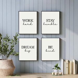 May include: Four framed art prints with black frames and white backgrounds. Each print features a different motivational phrase in black cursive font: "WORK hard", "STAY humble", "DREAM big", and "BE kind". The prints are displayed on a wooden surface.