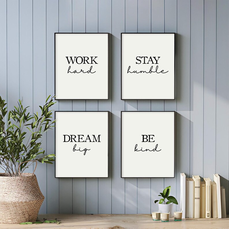 May include: Four framed art prints with black frames and white backgrounds. Each print features a different motivational phrase in black cursive font: "WORK hard", "STAY humble", "DREAM big", and "BE kind". The prints are displayed on a wooden surface.