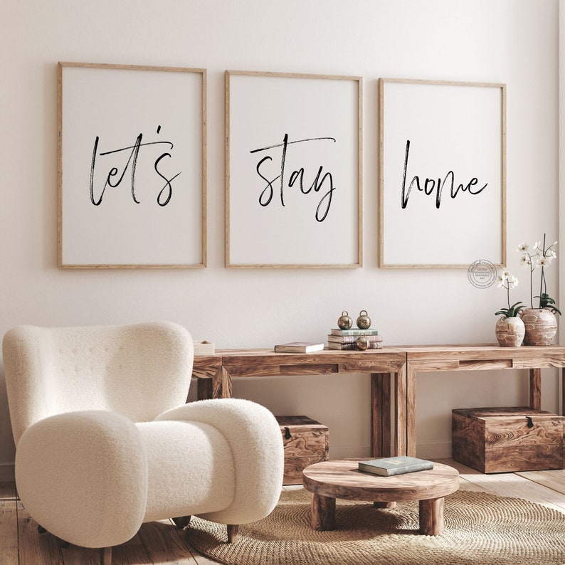 Pu&ograve; includere: Tre stampe incorniciate con il testo "Let's Stay Home" in scrittura nera su sfondo bianco. Le stampe sono appese a una parete bianca sopra un tavolo di legno e una poltrona bianca e soffice.