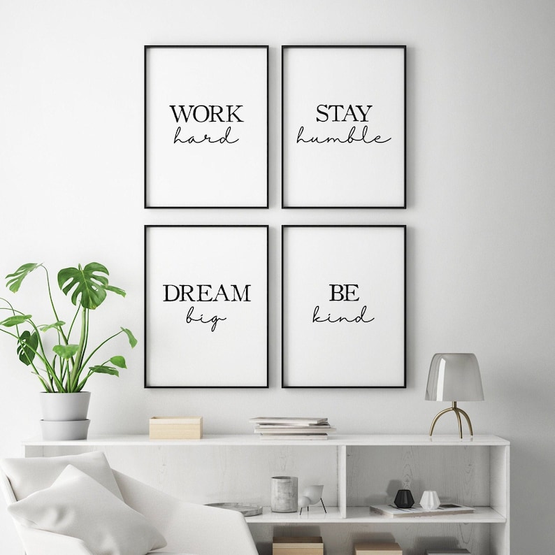 May include: Four framed art prints with black frames and white backgrounds. Each print features a motivational phrase in black script: "WORK hard", "STAY humble", "DREAM big", and "BE kind". A potted plant and a lamp are visible.