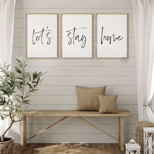 Pu&ograve; includere: Tre stampe incorniciate con le parole "Let's", "Stay" e "Home" in scrittura nera su sfondo bianco. Le stampe sono appese a una parete bianca a listelli di legno sopra una panca di legno con due cuscini marroni a motivi.