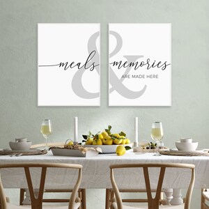 May include: Two white framed prints with a gray ampersand symbol and the text "meals & memories are made here" in black script font. The prints are hanging on a wall above a dining table set with white plates, glasses, and a bowl of lemons.