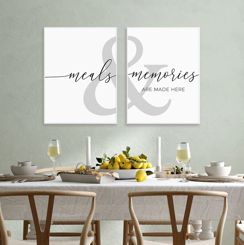 May include: Two white framed prints with a gray ampersand symbol and the text "meals & memories are made here" in black script font. The prints are hanging on a wall above a dining table set with white plates, glasses, and a bowl of lemons.