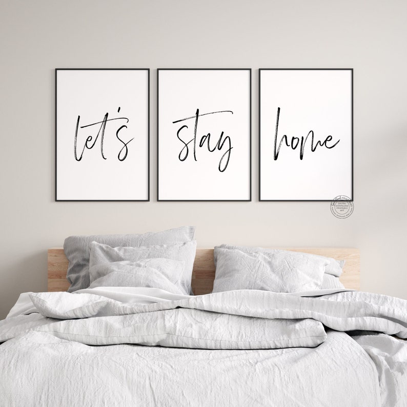 May include: Three black framed prints with white backgrounds and black calligraphy. The prints read "let's", "stay", and "home". The prints are hanging on a white wall above a bed with white bedding.