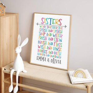 May include: A colorful framed print with the text "Sisters" and a list of activities, including "Splish and Splash", "Soap and Water", "Fresh and Clean", "Brush and Floss", "Wash and Rinse", "Bubbles and Bath", "Shampoo and Conditioner", and "Emma & Olivia".