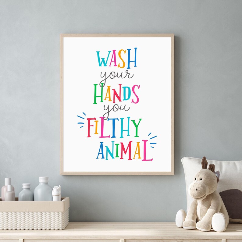 May include: A rectangular wall art print in a light wood frame featuring the text "WASH your HANDS you FILTHY ANIMAL" in a variety of bright, multi-colored letters. The words are stacked vertically against a plain white background.