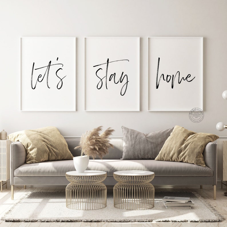 May include: Three black and white framed prints with the words "let's stay home" on a white background. The prints are hanging on a white wall above a gray couch with tan pillows and a vase of dried pampas grass.