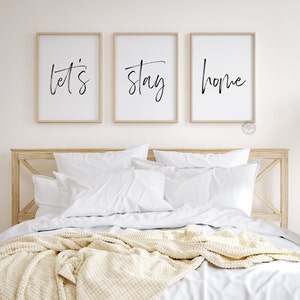 Pu&ograve; includere: Tre stampe incorniciate con le parole "Let's", "Stay" e "Home" in scrittura nera su sfondo bianco. Le stampe sono appese a un muro bianco sopra un letto con biancheria da letto bianca e una coperta di lana color crema.
