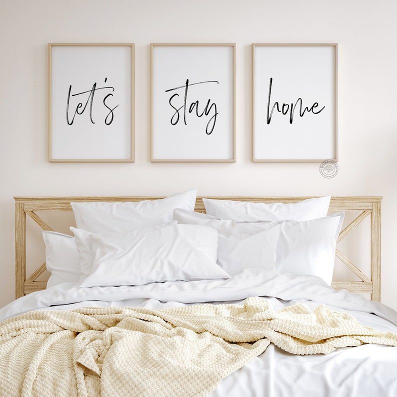 Pu&ograve; includere: Tre stampe incorniciate con le parole "Let's", "Stay" e "Home" in scrittura nera su sfondo bianco. Le stampe sono appese a un muro bianco sopra un letto con biancheria da letto bianca e una coperta di lana color crema.