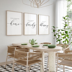 May include: Three framed prints with white backgrounds and black calligraphy text. The prints are hanging on a white wall above a wooden dining table with four chairs. The text on the prints reads "Having somewhere to go is home", "Having someone to love is family", and "Having both is a blessing".