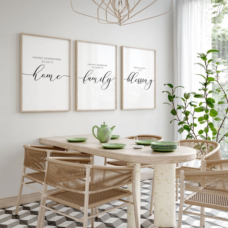 May include: Three framed prints with white backgrounds and black calligraphy text. The prints are hanging on a white wall above a wooden dining table with four chairs. The text on the prints reads "Having somewhere to go is home", "Having someone to love is family", and "Having both is a blessing".