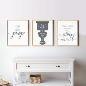 May include: Three framed wall art prints with light wood frames. The left print says "HAVE A NICE poop". The center print features a dark gray toilet graphic with the text "shit IS ABOUT TO GO DOWN". The right print says "WASH YOUR HANDS YOU filthy animal".