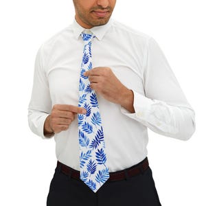 May include: A white tie with a blue leaf pattern. The tie is being adjusted by a person wearing a white dress shirt and black pants. The tie's design features a repeating pattern of blue leaves against a white background, creating a fresh and stylish look.