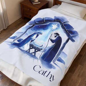 May include: A white fleece blanket featuring a blue watercolor illustration of a nativity scene with a star above, depicting figures gathered around a manger. The name Cathy is printed in dark blue script lettering at the bottom of the blanket.