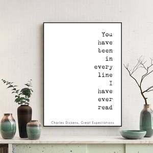 May include: A white framed print with a black and white quote from Charles Dickens' Great Expectations: "You have been in every line I have ever read". The print is displayed on a white shelf with a lamp, plants, and vases.