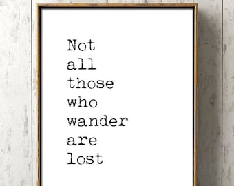 Puede incluir: Una impresión tipográfica en blanco y negro con la cita "Not all those who wander are lost". La impresión está enmarcada en un marco marrón simple y se muestra en un estante de madera azul claro.