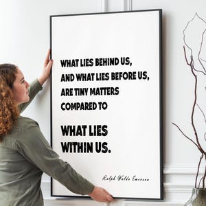 Puede incluir: Una impresi&oacute;n enmarcada con la cita "WHAT LIES BEHIND US, AND WHAT LIES BEFORE US, ARE TINY MATTERS COMPARED TO WHAT LIES WITHIN US." Una mujer cuelga el arte enmarcado en negro en una pared blanca. Un jarr&oacute;n y una mesa est&aacute;n en primer plano.