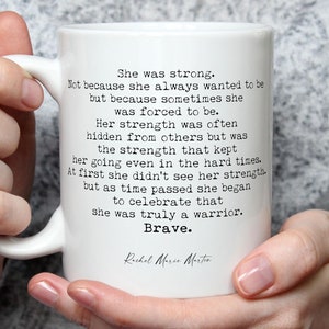 May include: White ceramic mug with a black inspirational quote about strength and bravery. The quote reads "She was strong. Not because she always wanted to be but because sometimes she was forced to be. Her strength was often hidden from others but was the strength that kept her going even in the hard times. At first she didn't see her strength but as time passed she began to celebrate that she was truly a warrior. Brave." The quote is attributed to Rachel More Morto.