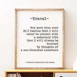 May include: A framed print with a quote about travel by Isabelle Eberhardt. The quote reads: "Now more than ever do I realize that I will never be content with a sedentary life that I will always be haunted by thoughts of a sun-drenched elsewhere." The print is hanging on a white wall above a wooden shelf with decorative items.