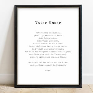 Vater Unser print in a black frame with a fine gold inner edge, leaning against a white wall on a light wood floor. The white print shows the title Vater Unser in bold typewriter lettering above the Lord's Prayer in German, centred, ending with Amen.
