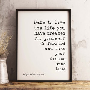May include: A black framed print with a white background and black text that reads 'Dare to live the life you have dreamed for yourself Go forward and make your dreams come true Ralph Waldo Emerson'. The print is sitting on a wooden surface with a silver pitcher and dried grass.
