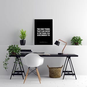 May include: A black framed print with white text that reads: "THE ONLY THING THAT CAN GROW IS THE THING YOU GIVE ENERGY TO" by Ralph Waldo Emerson. The print is displayed on a black desk with a white chair, plants, and a copper desk lamp.