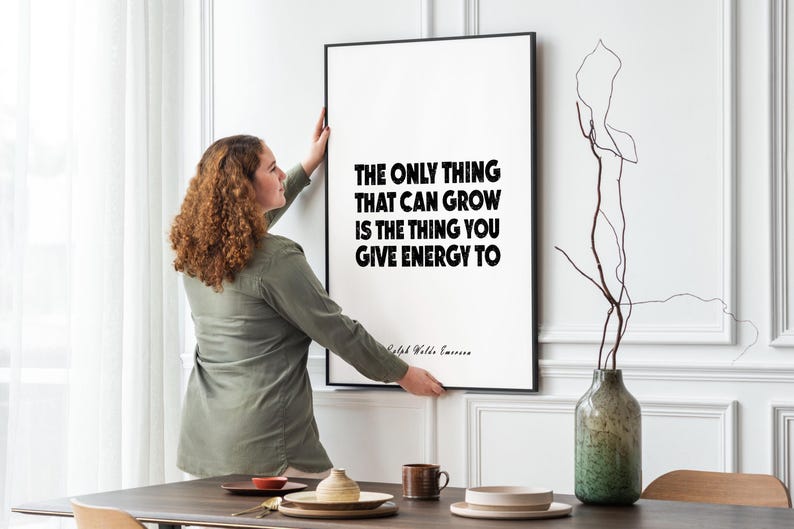 May include: A framed art print with the quote "THE ONLY THING THAT CAN GROW IS THE THING YOU GIVE ENERGY TO" in bold black text on a white background. The frame is black. A woman is hanging the art on a white wall.