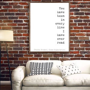 May include: A white framed print with a black and white quote from Charles Dickens' Great Expectations: "You have been in every line I have ever read". The print is hanging on a brick wall above a white couch with black and white patterned pillows.