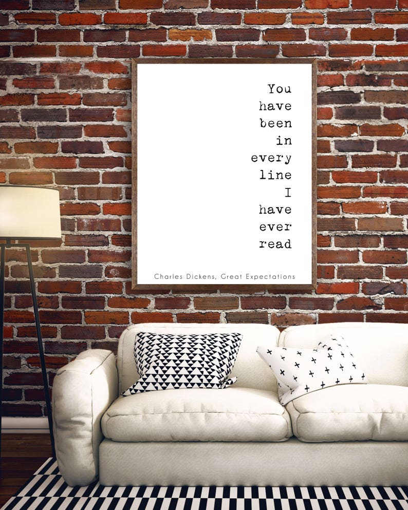 May include: A white framed print with a black and white quote from Charles Dickens' Great Expectations: "You have been in every line I have ever read". The print is hanging on a brick wall above a white couch with black and white patterned pillows.
