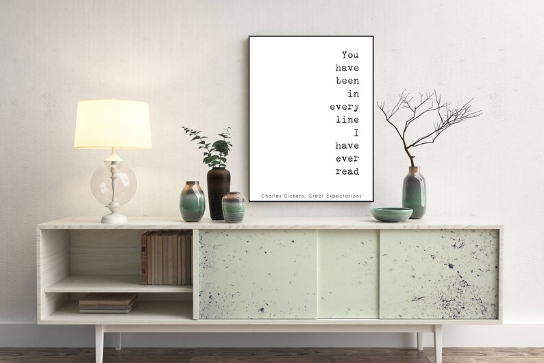 May include: A white framed print with a black and white quote from Charles Dickens' Great Expectations: "You have been in every line I have ever read". The print is displayed on a white shelf with a lamp, plants, and vases.