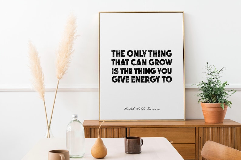 May include: A framed print with the quote "THE ONLY THING THAT CAN GROW IS THE THING YOU GIVE ENERGY TO" in black text. The print is in a gold frame, and is on a wooden cabinet. A small potted plant and decorative items are nearby.