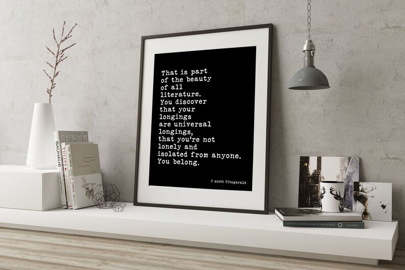 May include: A framed wall art print featuring a black background with white typewriter-style text that reads: That is part of the beauty of all literature. You discover that your longings are universal longings, that you're not lonely and isolated from anyone. You belong. F. Scott Fitzgerald.