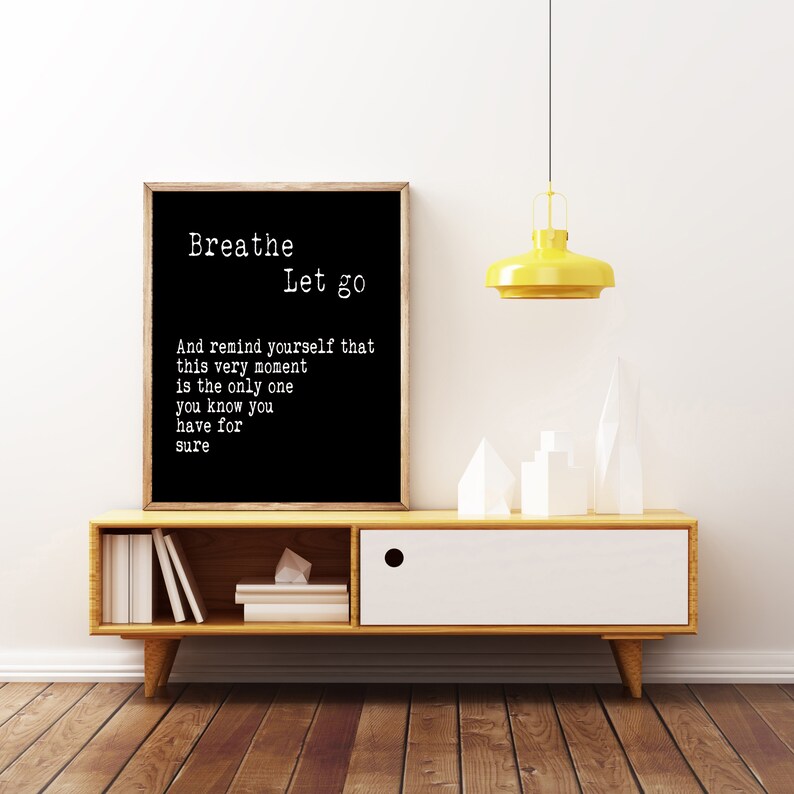 May include: Black and white typography print with the text "Breathe Let go" and a quote about reminding yourself that this moment is the only one you know for sure. The print is framed and hanging on a white wall above a wooden shelf with a white drawer.