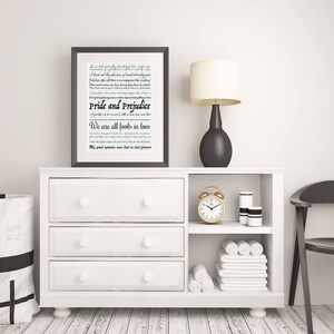 May include: A white dresser with three drawers and two shelves. The top shelf has a black framed print with the text "Pride and Prejudice" and a quote from the book. A black lamp with a white shade sits on the dresser. A gold alarm clock and two white candles are on the lower shelf. White towels are stacked on the bottom shelf.