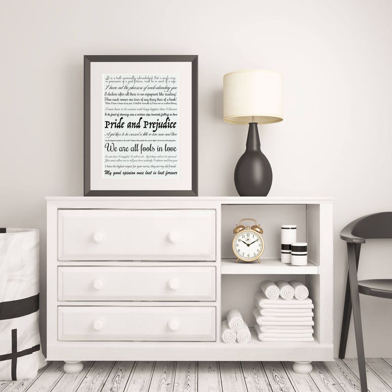 May include: A white dresser with three drawers and two shelves. The top shelf has a black framed print with the text "Pride and Prejudice" and a quote from the book. A black lamp with a white shade sits on the dresser. A gold alarm clock and two white candles are on the lower shelf. White towels are stacked on the bottom shelf.