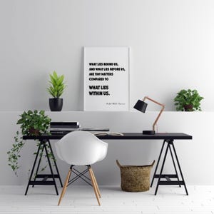 Puede incluir: Una moderna configuraci&oacute;n de oficina en casa con un escritorio negro, una silla blanca y una impresi&oacute;n enmarcada con el texto "WHAT LIES BEHIND US...WHAT LIES WITHIN US." Una l&aacute;mpara de escritorio de cobre y negra, plantas en macetas y una cesta de mimbre completan la decoraci&oacute;n.