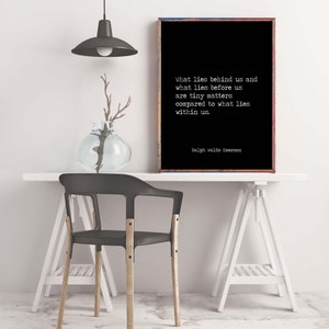 May include: Black and white typography print with a quote by Ralph Waldo Emerson: "What lies behind us and what lies before us are tiny matters compared to what lies within us." The print is framed and hanging on a white wall above a white table with a black chair.