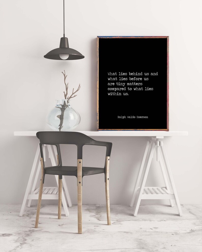 May include: Black and white typography print with a quote by Ralph Waldo Emerson: "What lies behind us and what lies before us are tiny matters compared to what lies within us." The print is framed and hanging on a white wall above a white table with a black chair.