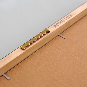 May include: Close-up of a wooden picture frame with a gold-colored sawtooth hanger and the text "ADHO101" printed on the back.
