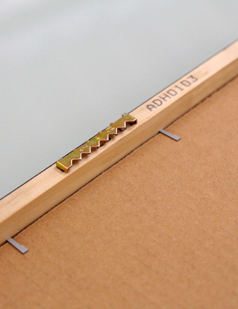 May include: Close-up of a wooden picture frame with a gold-colored sawtooth hanger and the text "ADHO101" printed on the back.